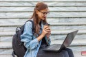 Happy modern woman student in denim jacket and backpack sitting on the stairs with laptop outdoor. Drink coffee. Watching video. Distance learning. Modern youth concept.